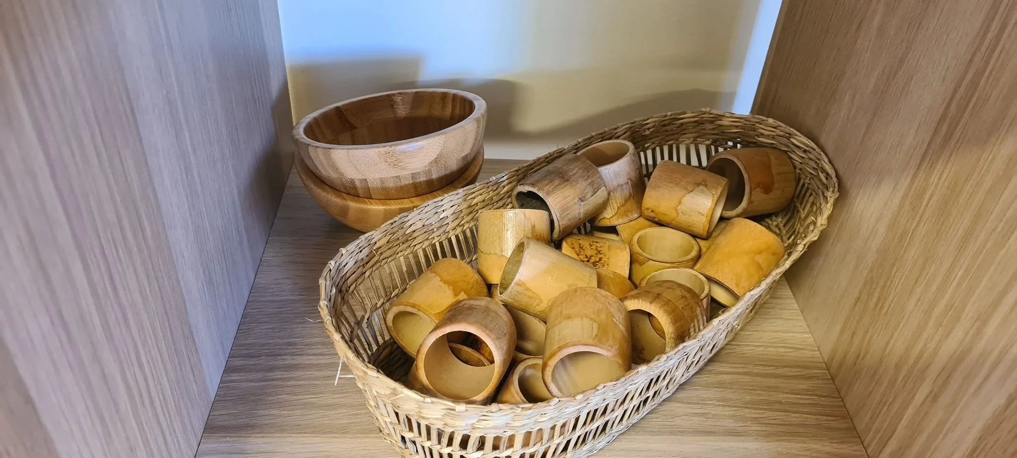 A wicker basket filled with small wooden cups and a wooden bowls on a wooden surface in a corner.