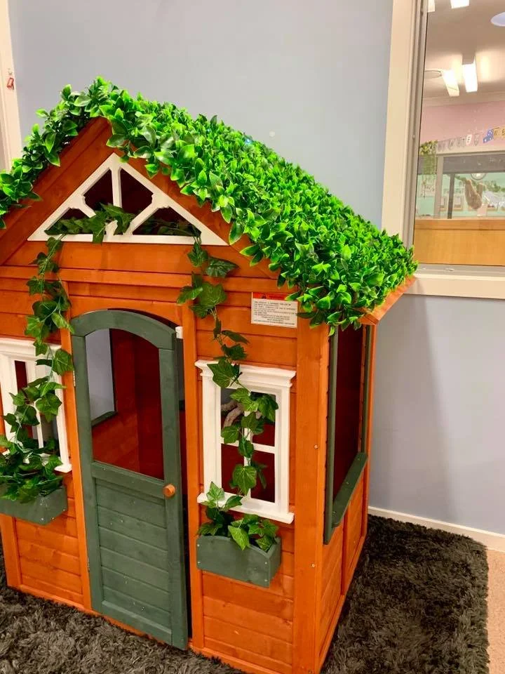 A small wooden playhouse with a green door, white-trimmed window, and a faux ivy vine climbing up its side, situated indoors on a plush carpet.