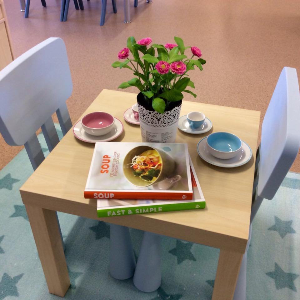 A small wooden table with a potted pink flower plant, two sets of pastel-colored bowls and saucers, and two cookbooks titled 'Soup' and 'Fast & Simple' in a child's or cozy space, with a star-patterned rug underneath.
