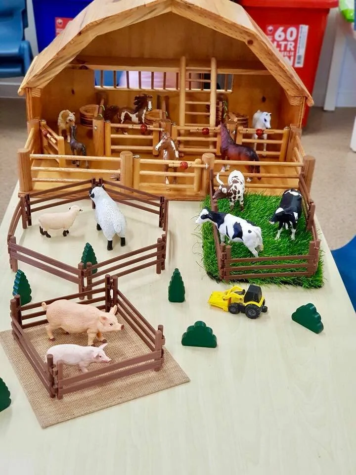A miniature farm playset with a barn, various small toy animals including pigs, cows, sheep, horses, and chickens, a toy tractor, and small trees on a table.