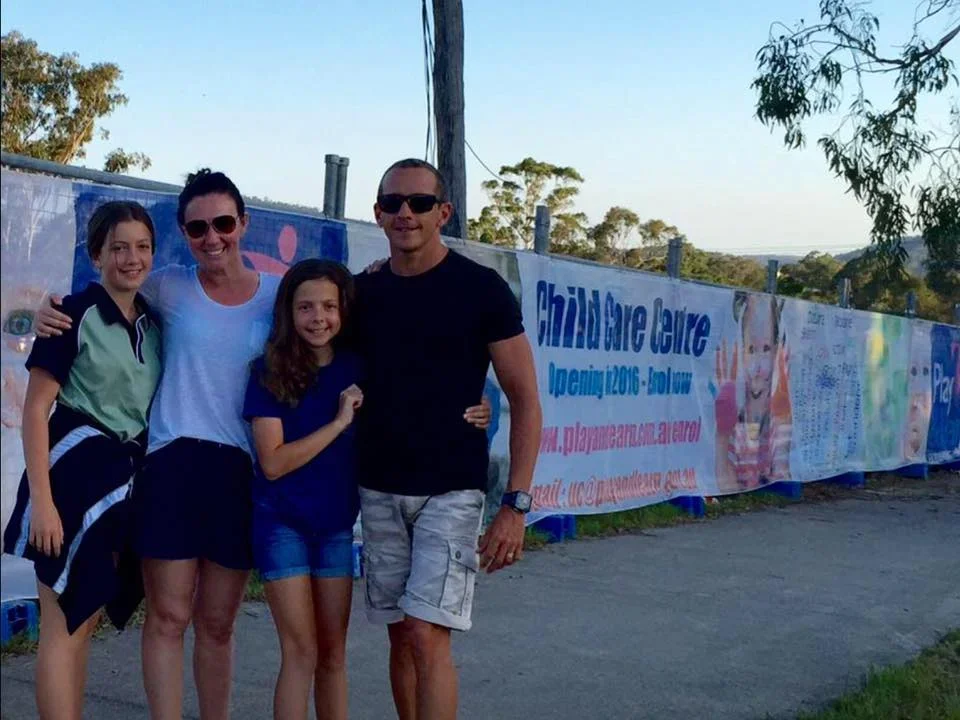 Four people, two adults and two children, standing outdoors near a colorful billboard with trees and a clear sky in the background.
