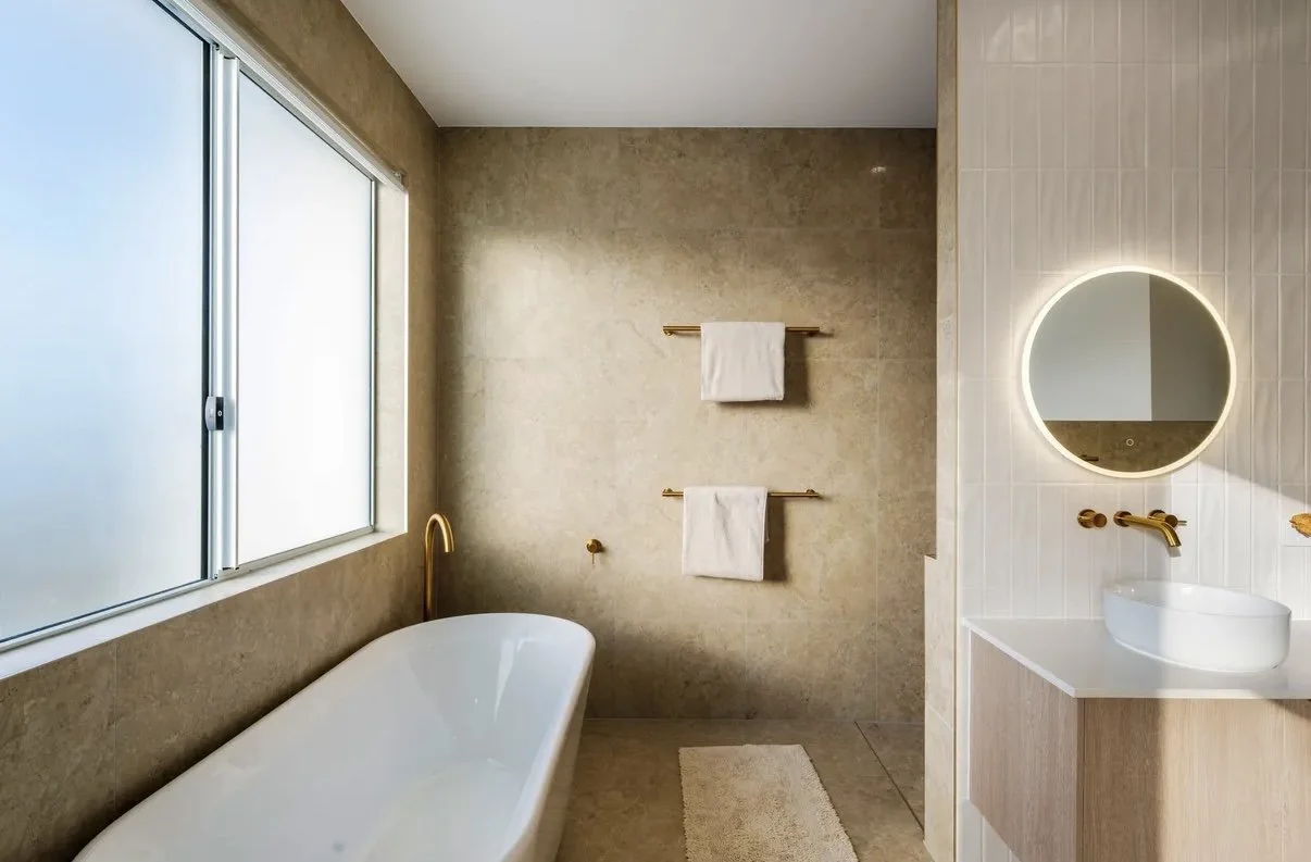 Modern bathroom with a large window, white bathtub with gold fixtures, beige walls, a white vanity with a vessel sink, a round mirror with backlight, white towels on gold towel racks, and a small beige rug.