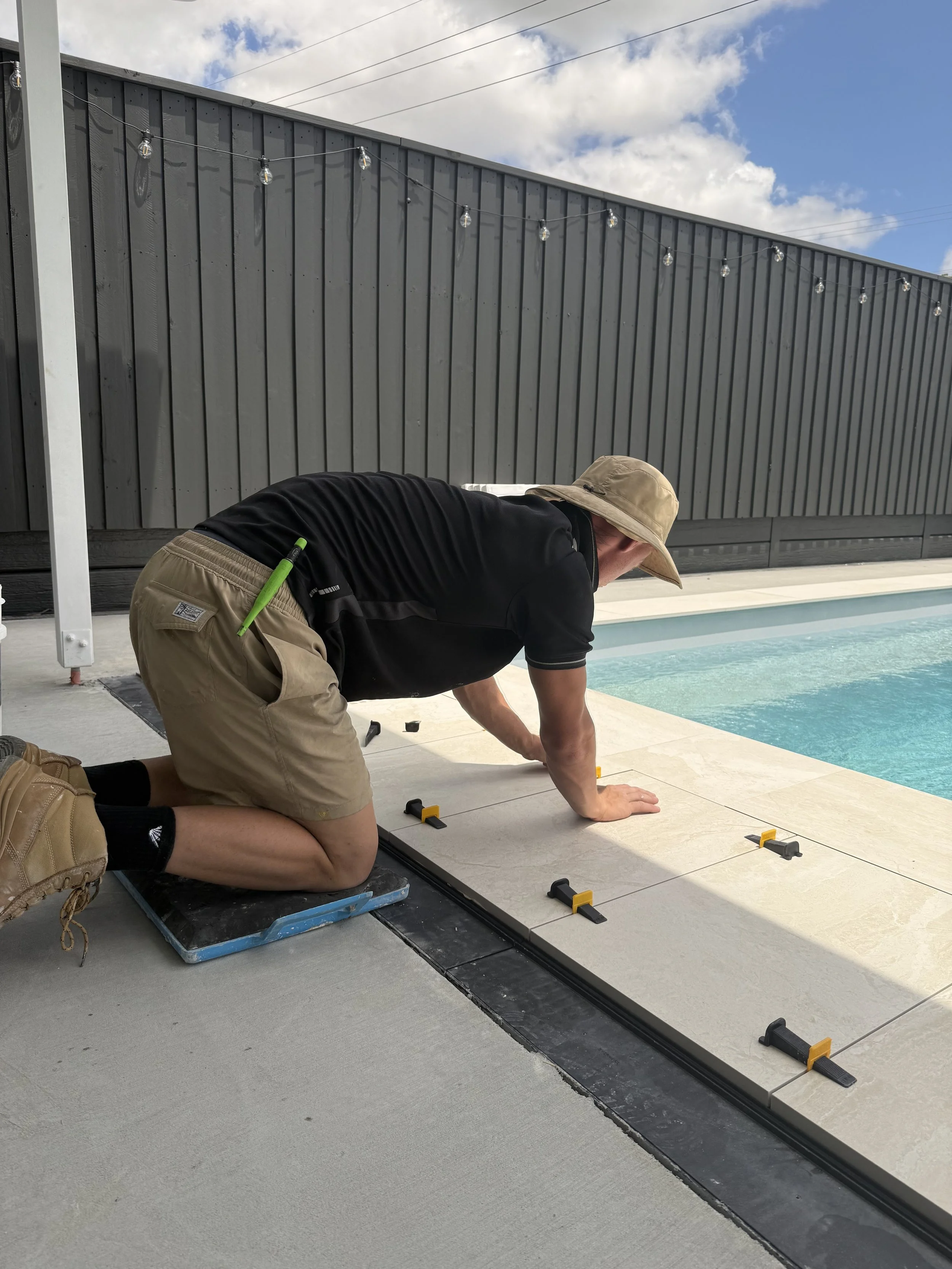 Person working on tile installation near a swimming pool, with a gray privacy fence and string lights in the background.