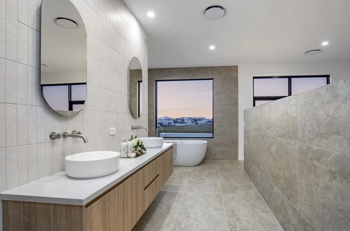 Modern bathroom with two vessel sinks, oval mirrors, wooden vanity, bathtub, large window showing a sunset view, tiled walls and floors, ceiling lights, and a partially partitioned area.