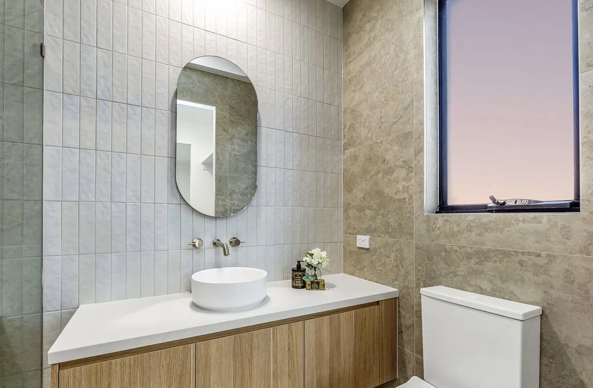 Modern bathroom with a round vessel sink, a wooden vanity, a small mirror above the sink, a toilet, a window with a blind, and decorative items on the countertop.