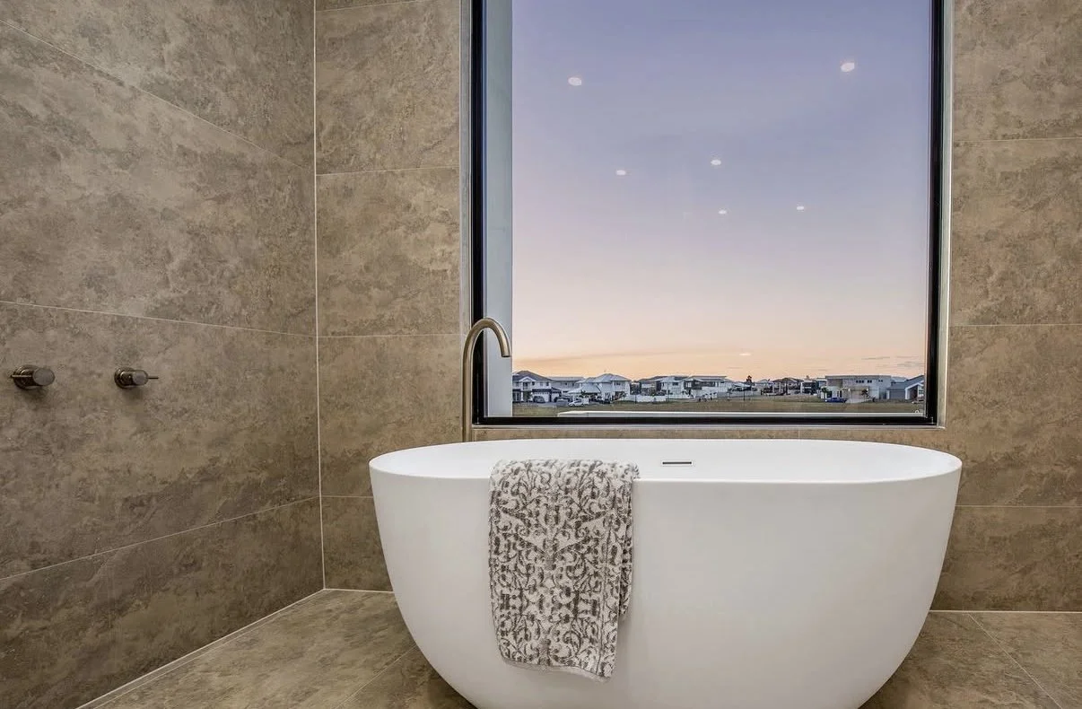 Modern bathroom with a freestanding bathtub in front of a large window showing a sunset view of houses outside.