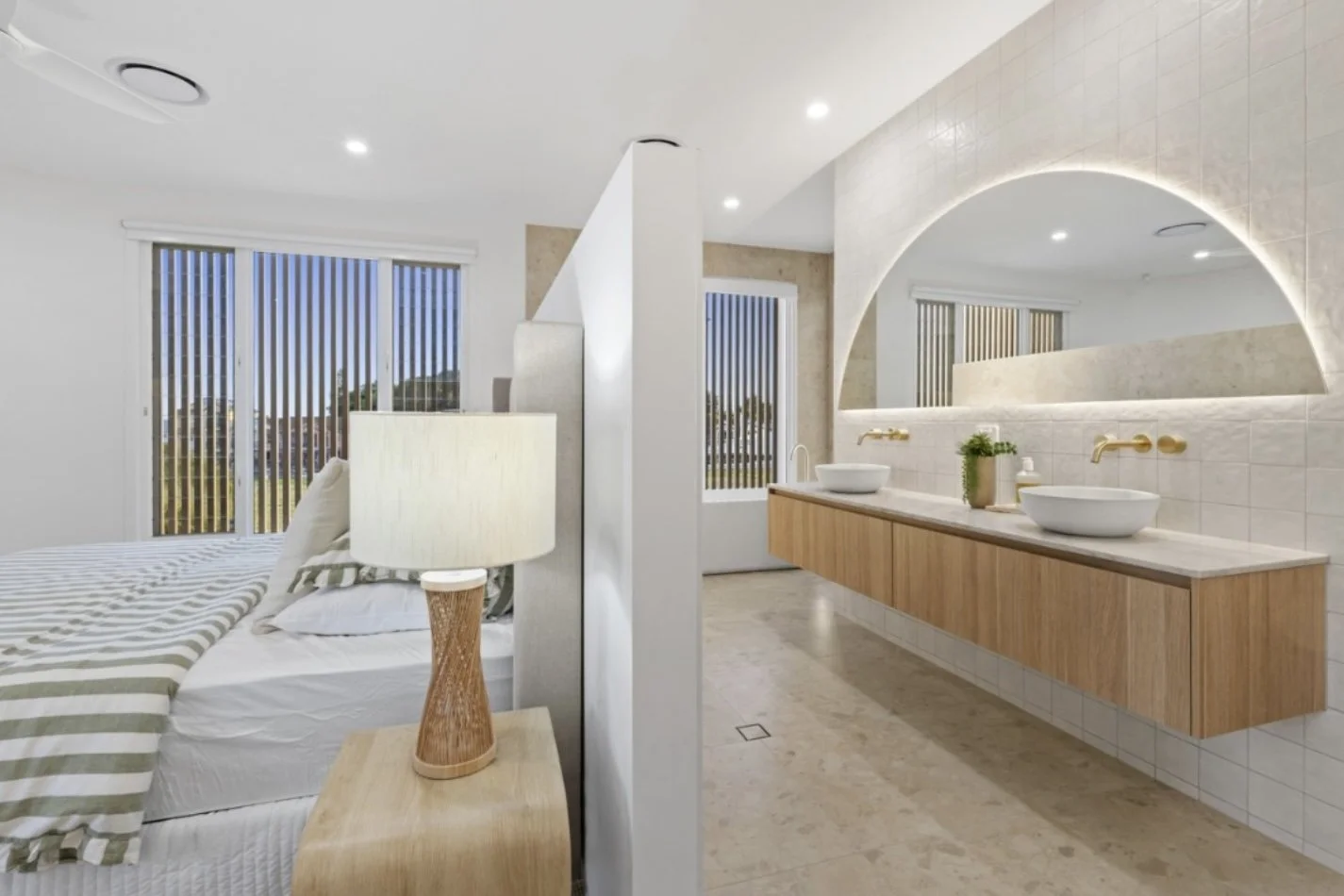 Modern bedroom with a bed, lamp, and large sliding glass doors, connected to a bathroom with two vessel sinks, a large mirror, and a wooden vanity.
