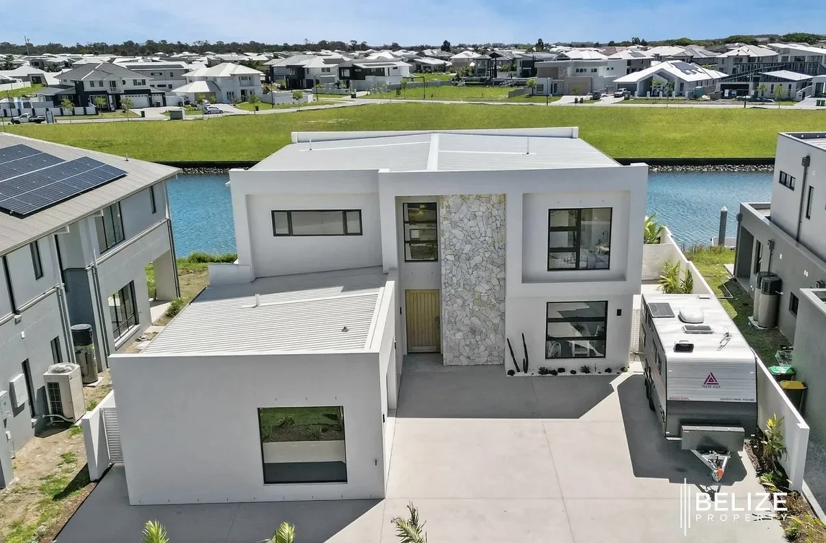 Modern white house with geometric design, stone accent wall, and flat roof, situated near water with neighboring houses and a trailer parked in the driveway.