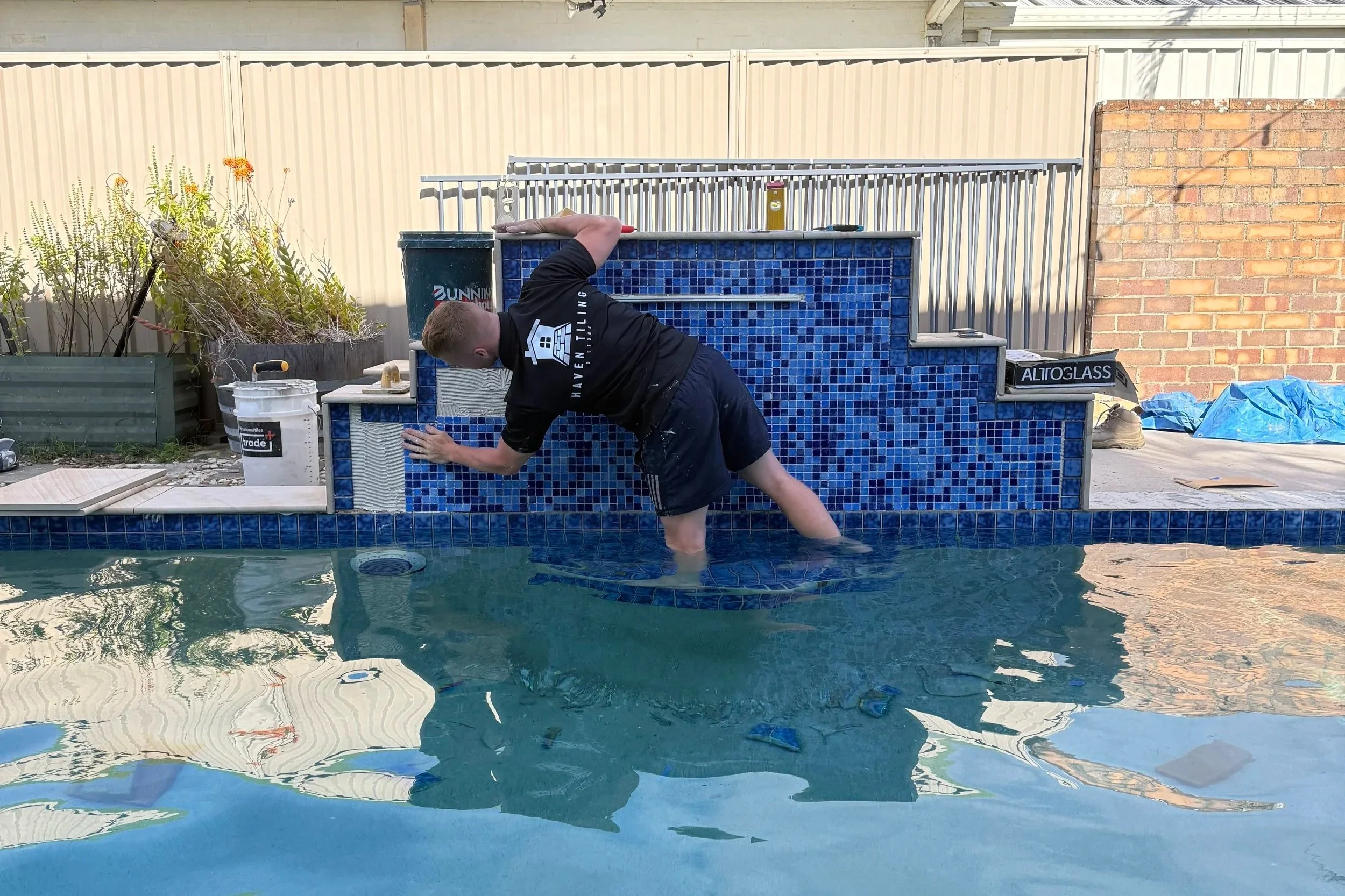 Person working on a house project by a backyard pool, with blue tiled wall, gardening tools, and a bucket nearby.
