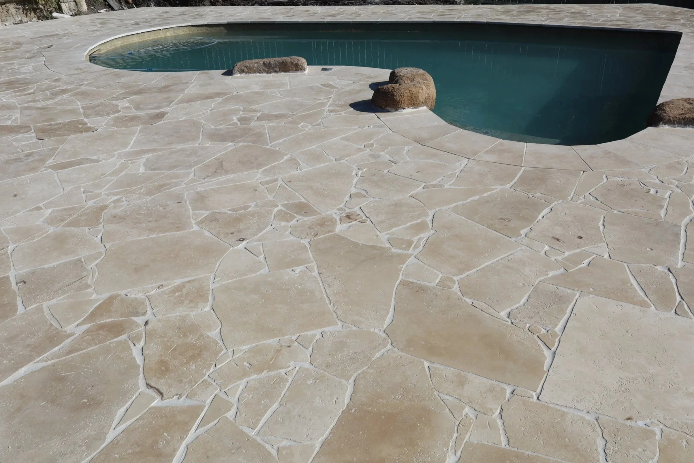 A kidney shaped backyard pool with turquoise water, surrounded by natural stone crazy pave completed by Haven Tiling & Stone Pty Ltd tilers, feature rocks are also placed around the pool coping tiles to create a more natural and luxury designed pool.