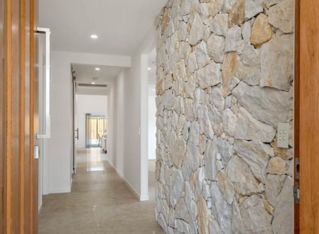 Interior of a modern house hallway with a stone accent wall on the right, white walls, wooden trim, and door frames, with natural light coming in from a door at the end of the hall.