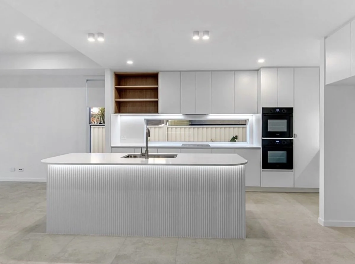 Modern white kitchen with island, built-in ovens, wooden shelves, and a window.