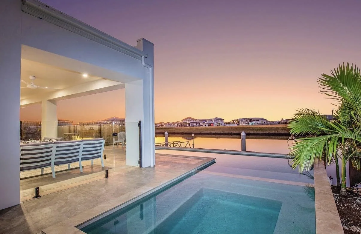 An outdoor pool area at sunset with a view of a canal and houses in the distance. The area is part of a modern house with a patio and glass sliding doors, and there's a green palm plant on the right side.
