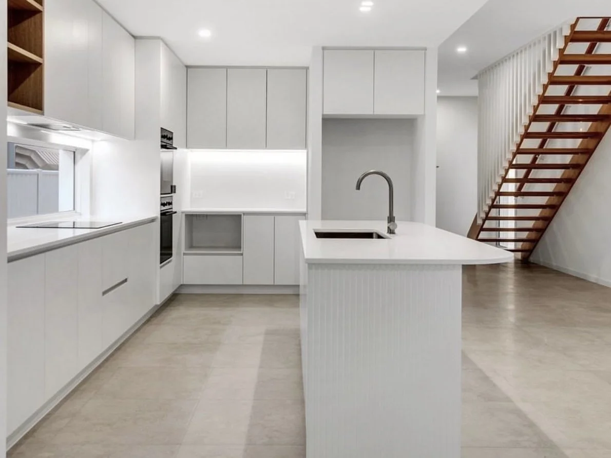 Modern white kitchen with island, built-in oven, and staircase with wooden steps.