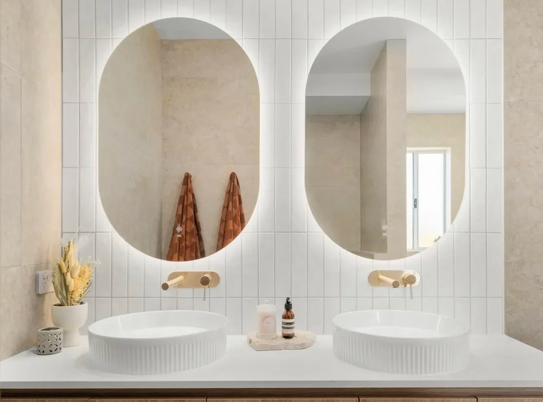 Bathroom double vanity with two white vessel sinks, wall-mounted gold faucets, large mirrors, beige tiled walls, and decor including a plant, soap, and towel.