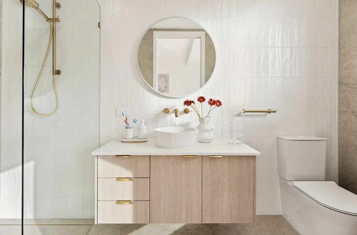 Modern bathroom with white tiled walls, a wooden vanity with gold handles, a white countertop, a round mirror, a flower vase with red flowers, a toothbrush holder, a soap dispenser, a glass container, and a toilet. There is also a walk-in shower with