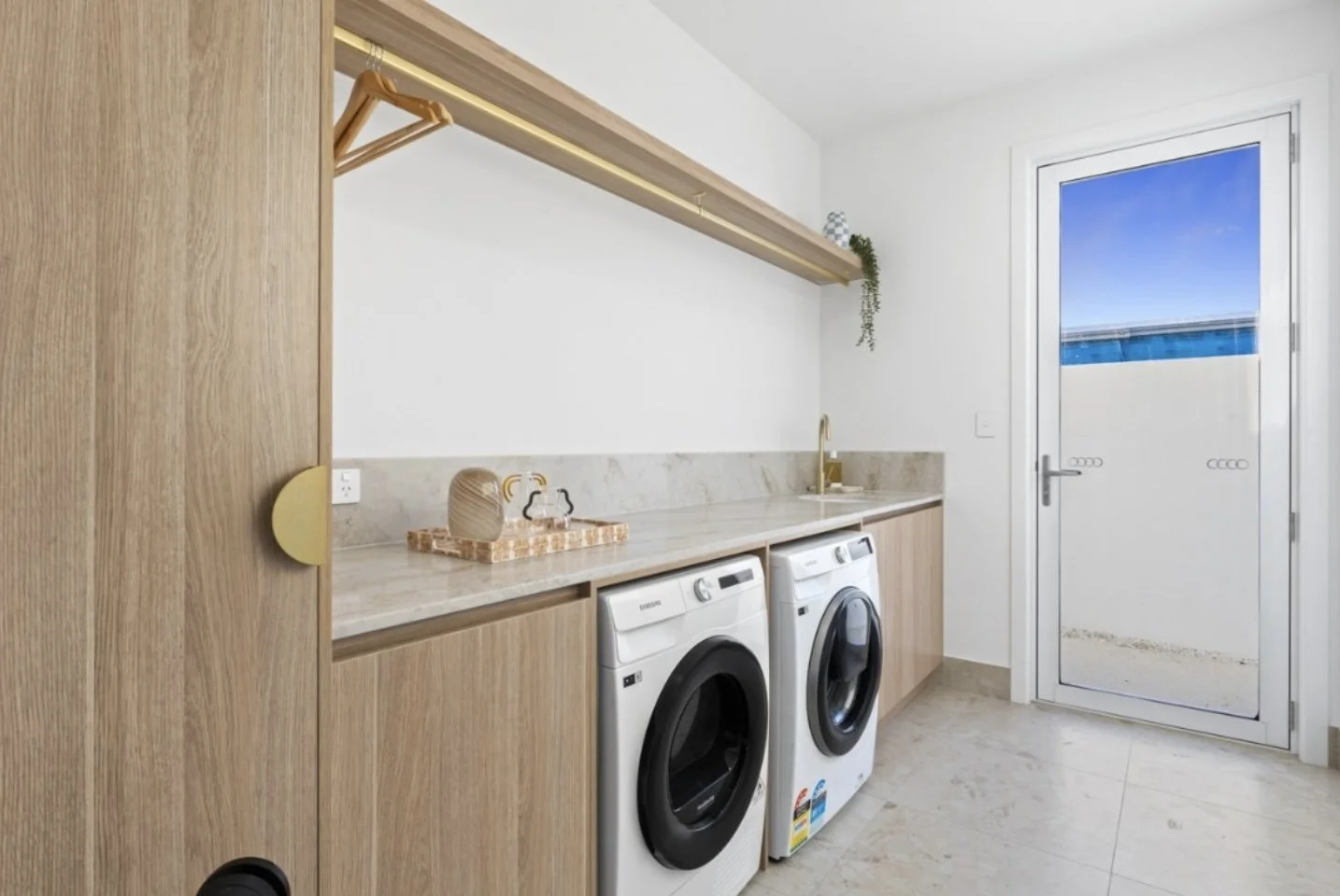 A laundry room with a wooden cabinet and a countertop, two washing machines, a door leading outside to a balcony, and minimal decor including a hanging plant and decorative pieces on the countertop.