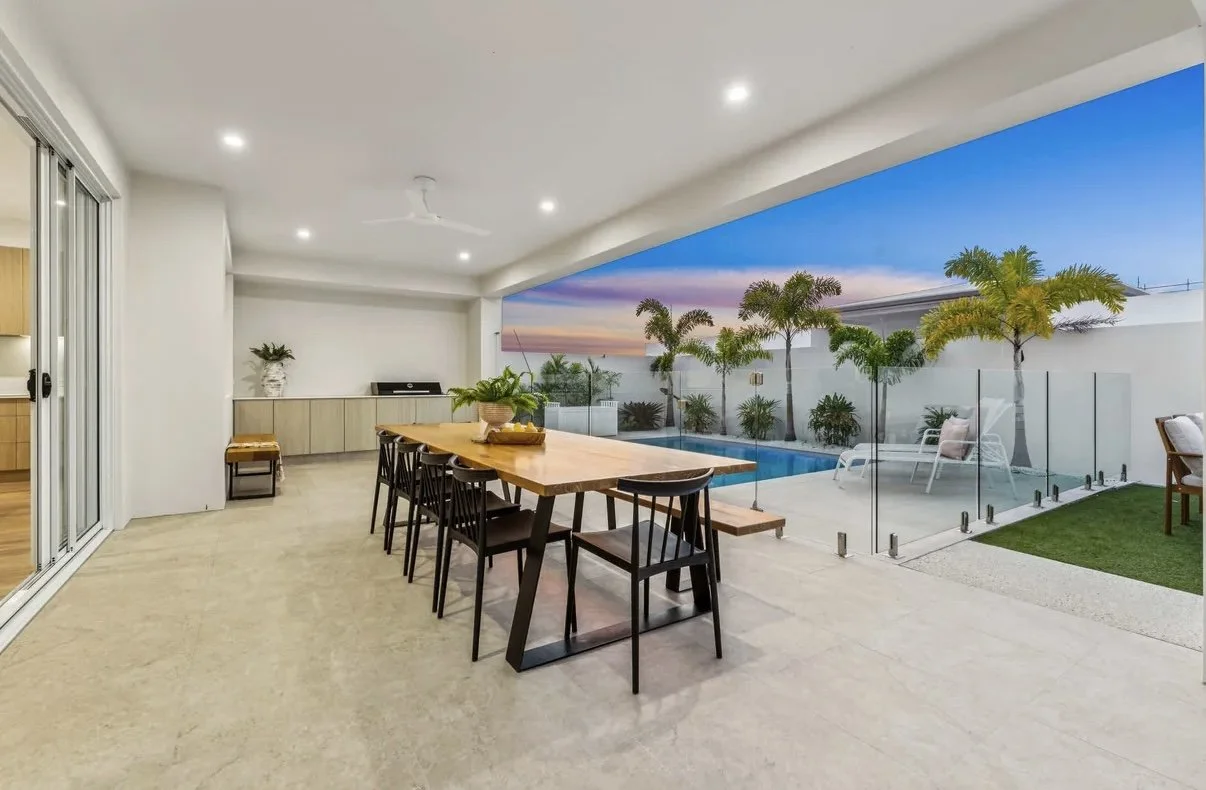 Open patio with dining table and chairs, glass fence, outdoor pool, lounge chairs, palm trees, and sunset sky.