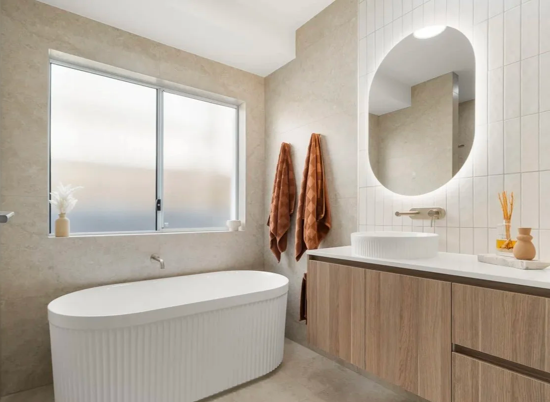 Modern bathroom with a large frosted window, a white freestanding bathtub, wooden vanity with a white vessel sink, a round mirror, and two brown towels hanging on the wall.