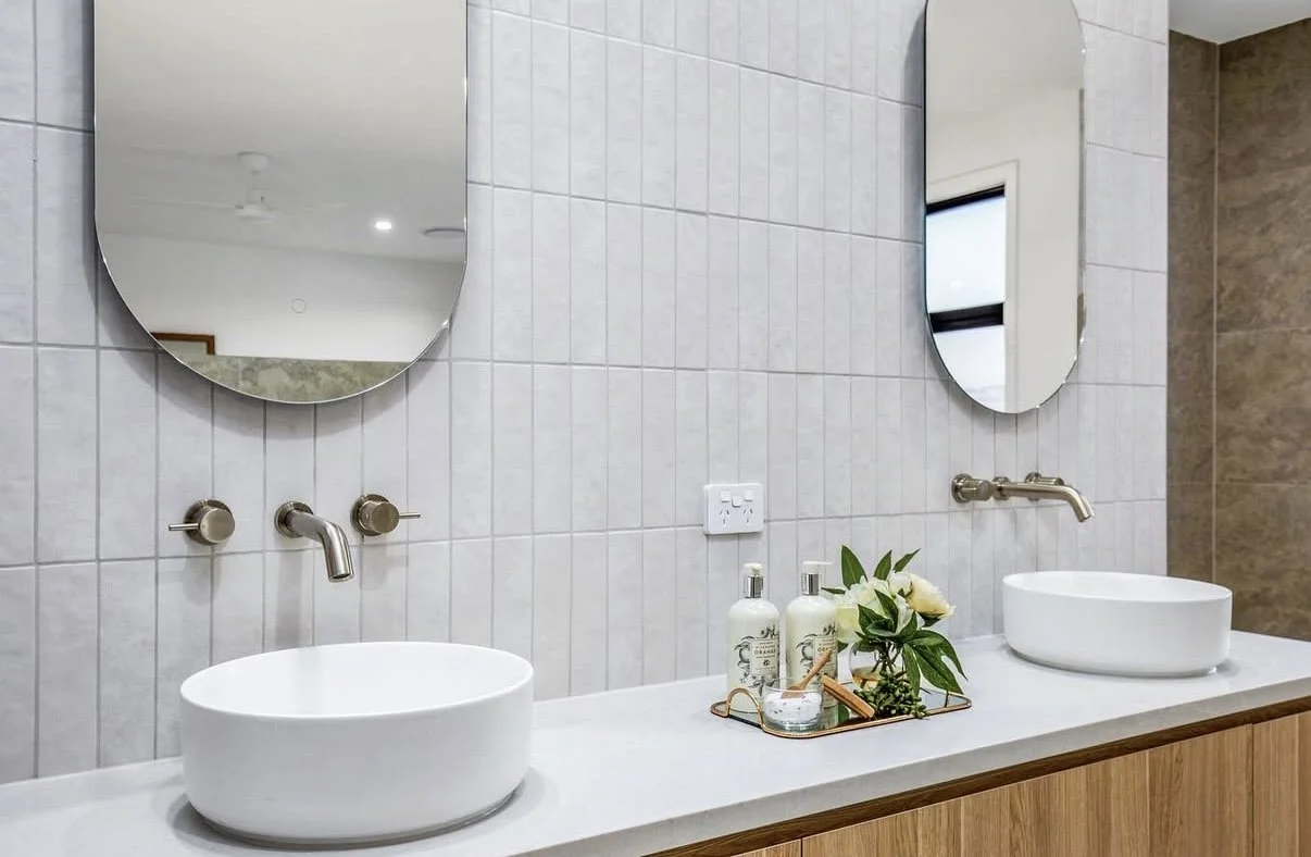 Bathroom with two white oval sinks, two wall-mounted faucets, two mirrors, a wooden vanity, and a tray with soap, lotion, a plant, and glasses.