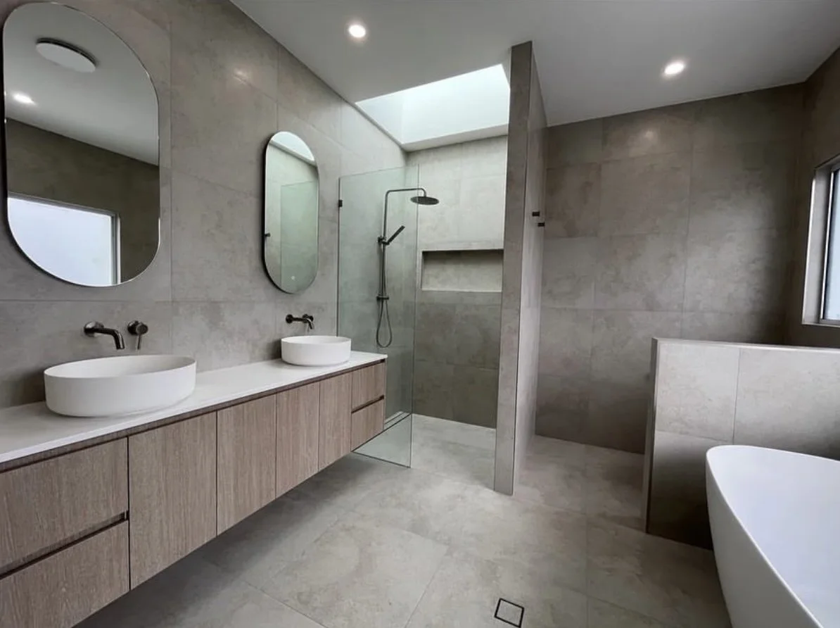 Modern bathroom with dual sinks, oval mirrors, a walk-in shower with a skylight, and a clawfoot bathtub, with neutral gray tones and minimalist decor.