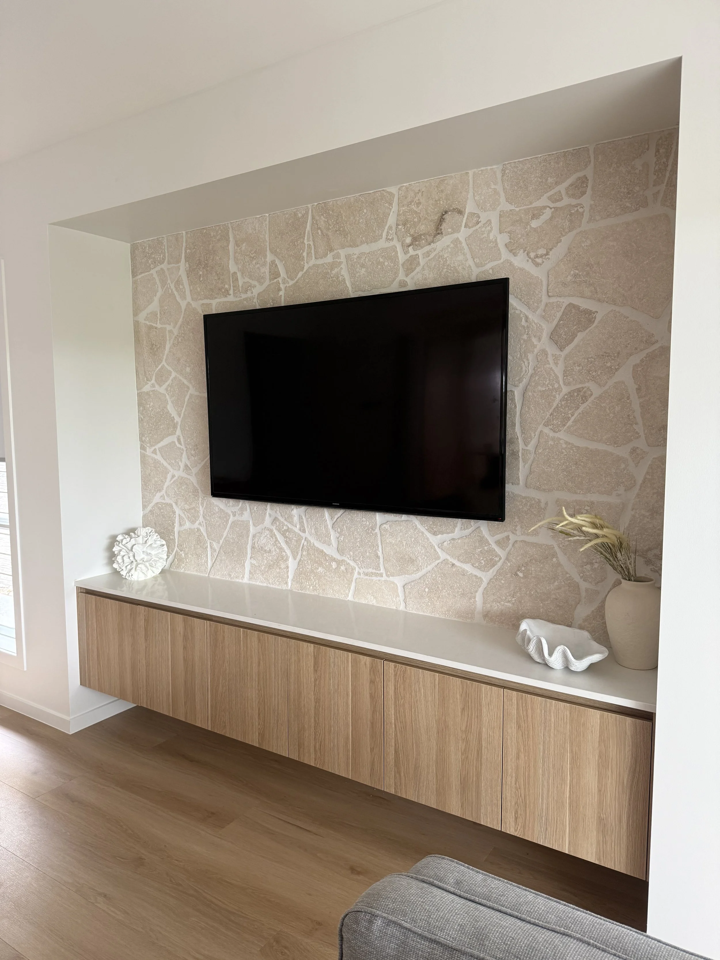 Living room television wall with beige stone accent wall, mounted flat-screen TV, wooden floating cabinet, and decorative items including a white shell sculpture, a beige vase with dried plants, and a white decorative shell on the cabinet.