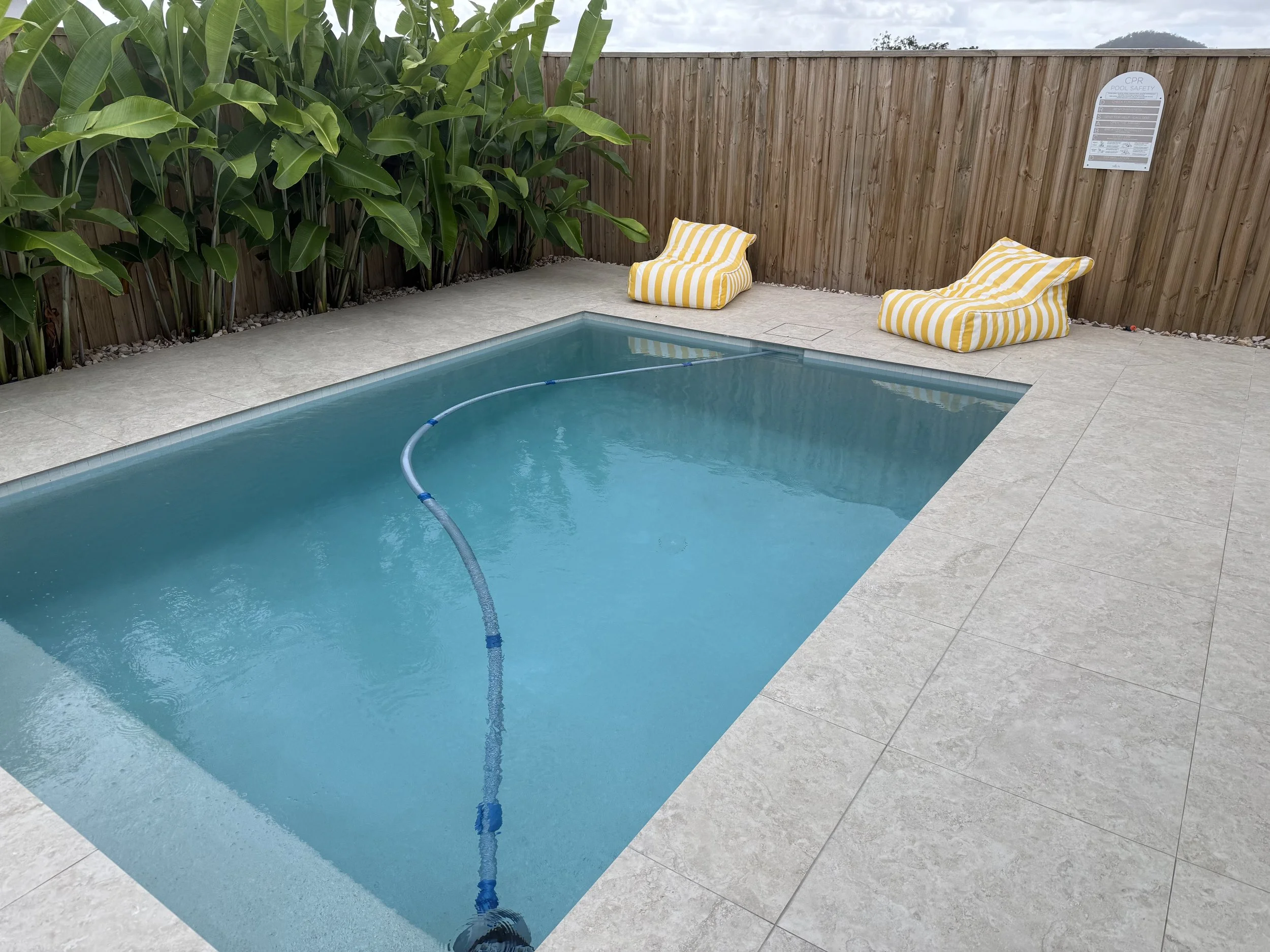 A small backyard pool with turquoise water, surrounded by a light-colored tile patio, wooden privacy fence, and green tropical plants; two yellow and white striped pool lounge chairs are positioned on the patio.