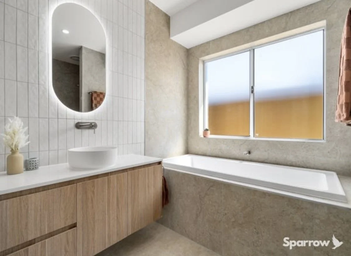 Modern bathroom with a large window, a bathtub, a wooden vanity with a vessel sink, an oval mirror, and decorative items on the counter.