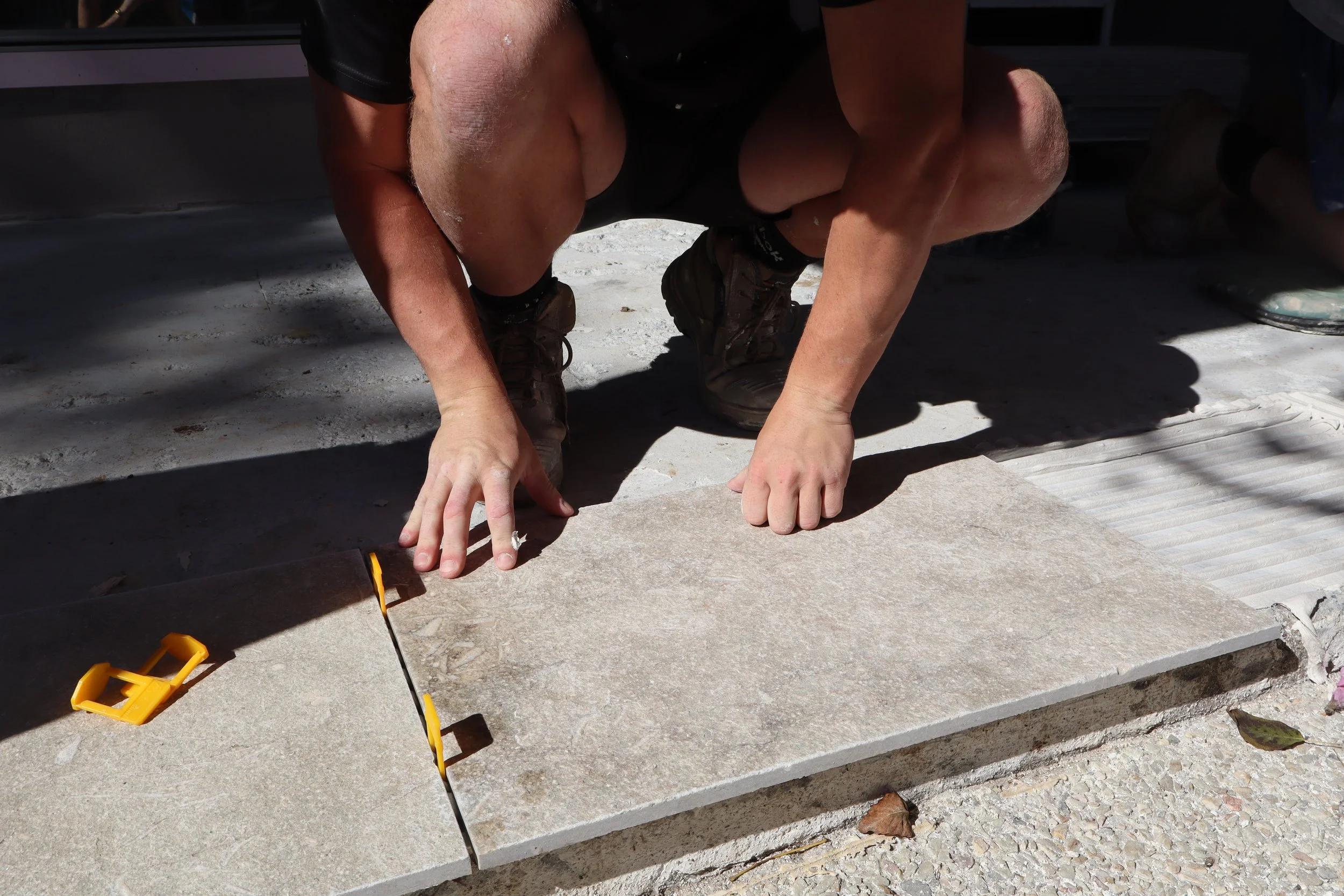 Person crouching on a concrete surface, laying a 20mm thick tile with construction and tiling tools nearby.