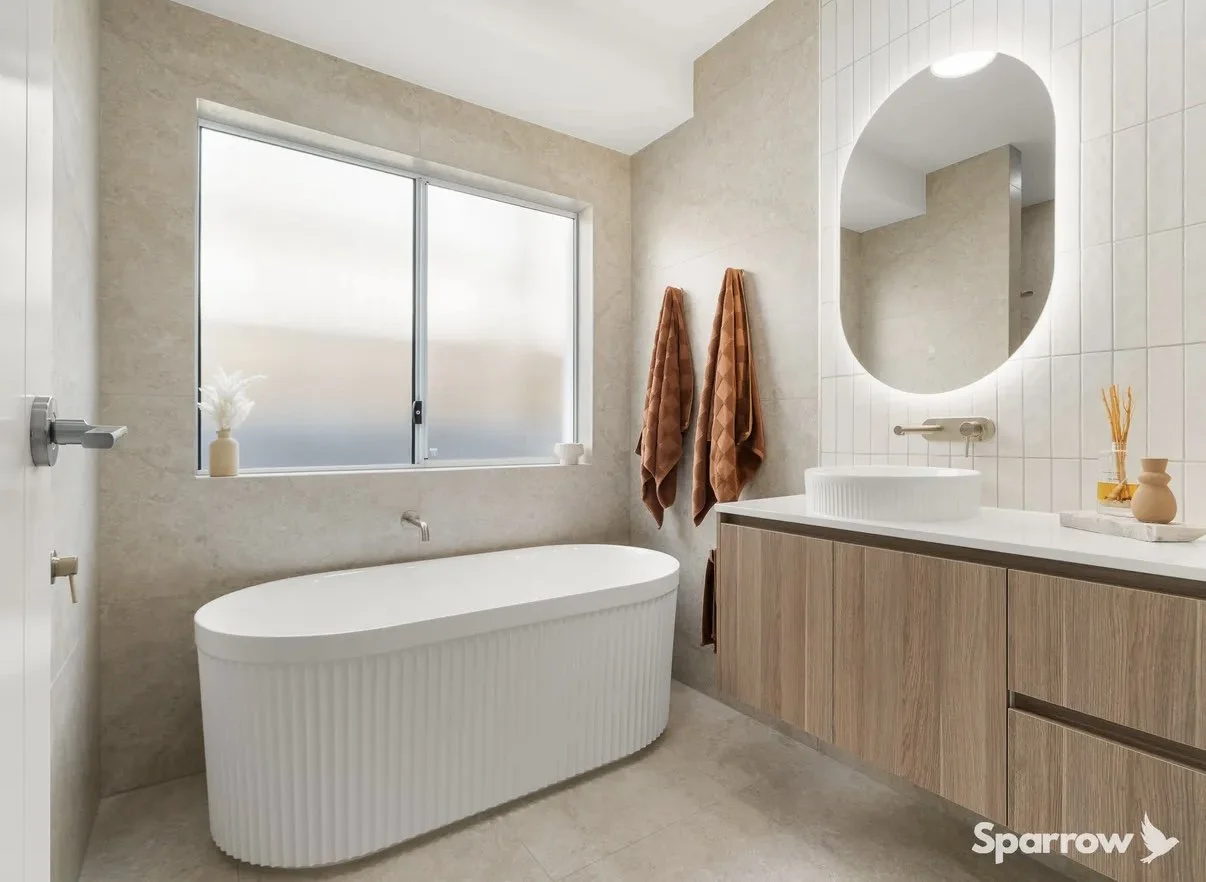 Modern bathroom with a freestanding bathtub under a large frosted window, wooden vanity with a round vessel sink, oval mirror, and orange towels hanging on the wall.
