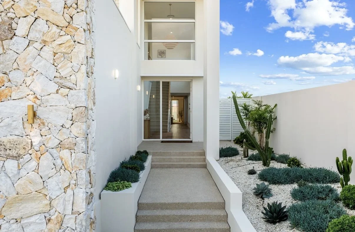 Modern house entrance with stone accent wall on the left, glass sliding door, and desert-style landscaping with succulents and cacti on the right under a blue sky with clouds.