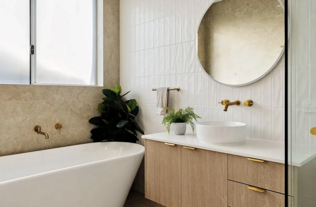 Modern bathroom with a white freestanding bathtub, a wooden vanity with a vessel sink, a large round mirror, gold fixtures, a small towel on a gold towel bar, a green plant, and a window with frosted glass.