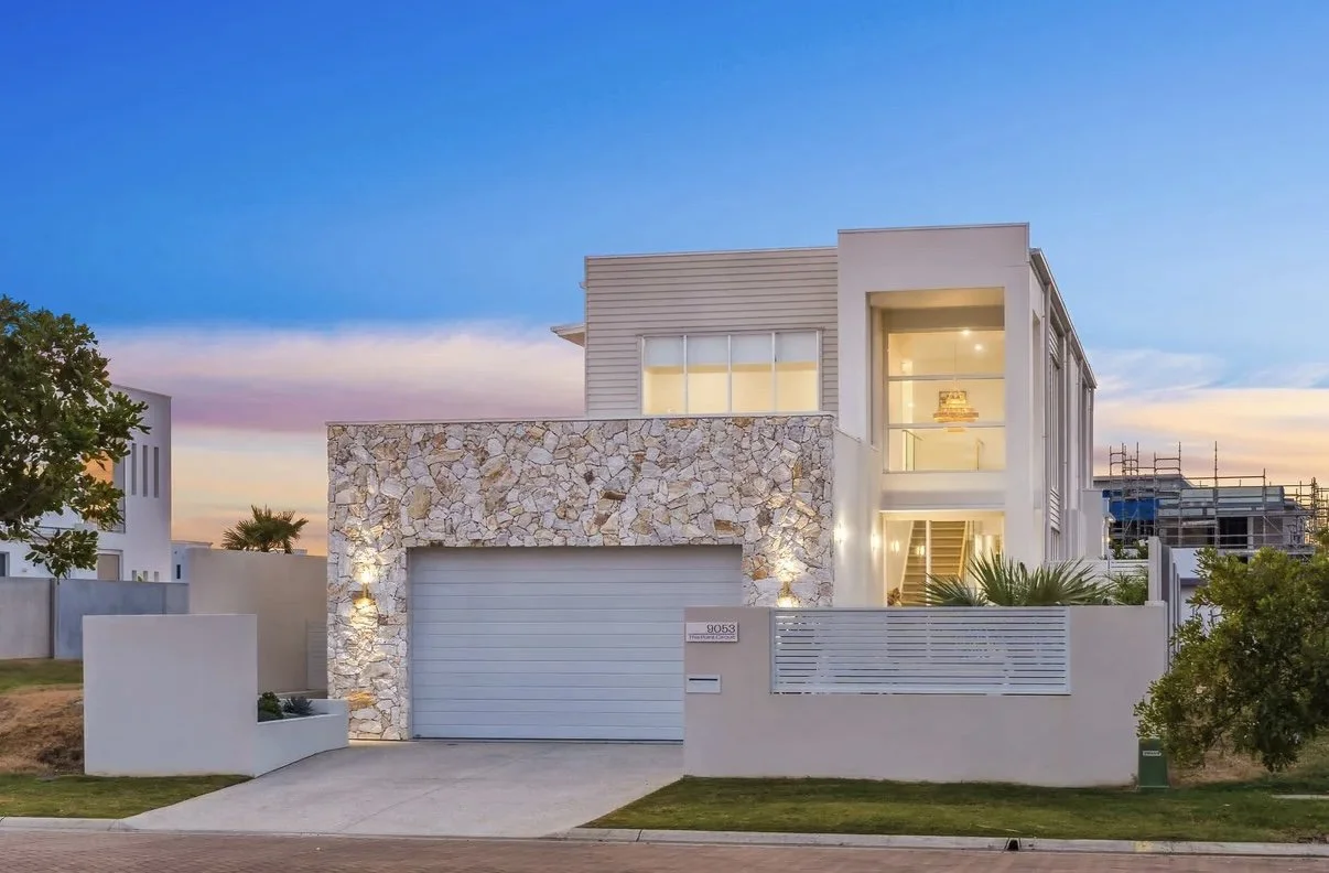 Modern two-story house with a stone facade, large windows, and a garage, surrounded by a low wall and landscaping, at sunset.