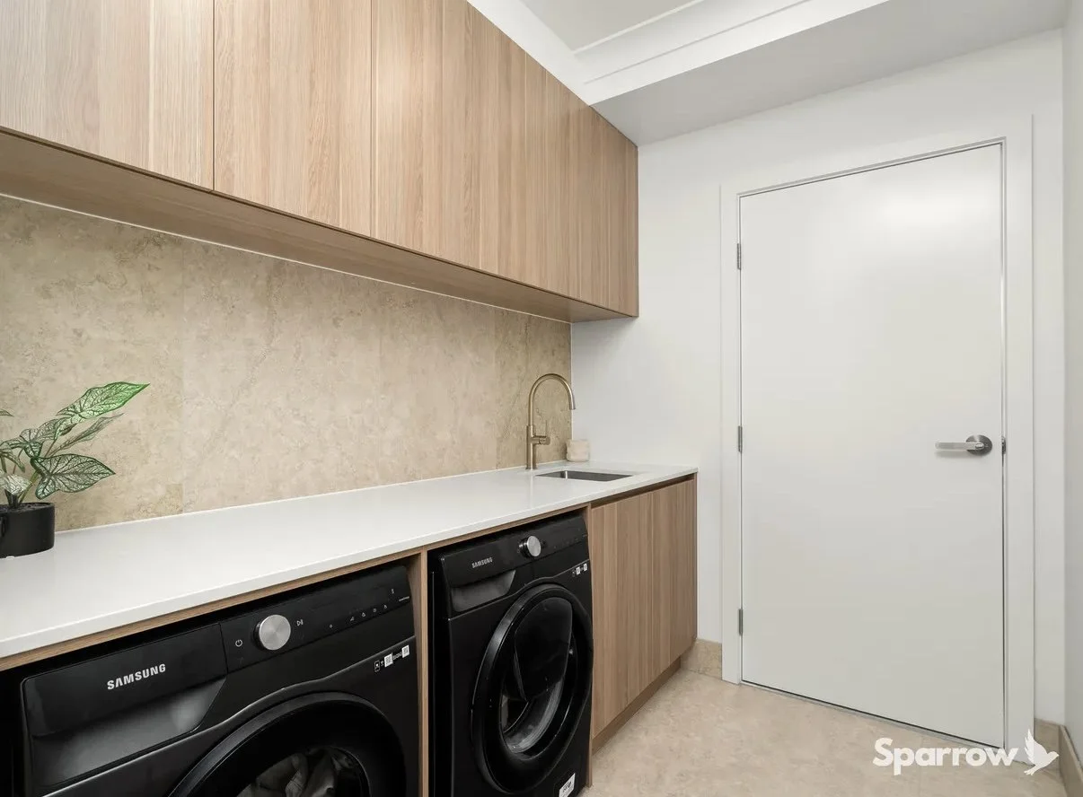 Laundry room with wooden cabinets, a white countertop, a small sink with a gold faucet, and a washing machine. There is a wall plant on the left and a closed white door on the right.