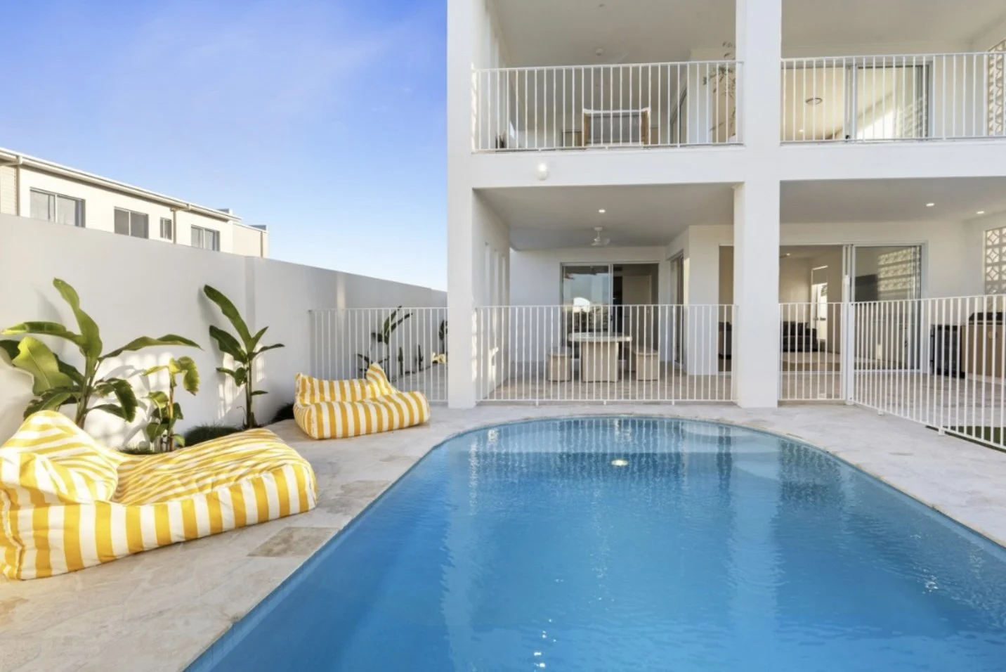 Backyard with a swimming pool, yellow and white striped cushions, and potted plants, enclosed by a white fence, with a white modern building in the background.