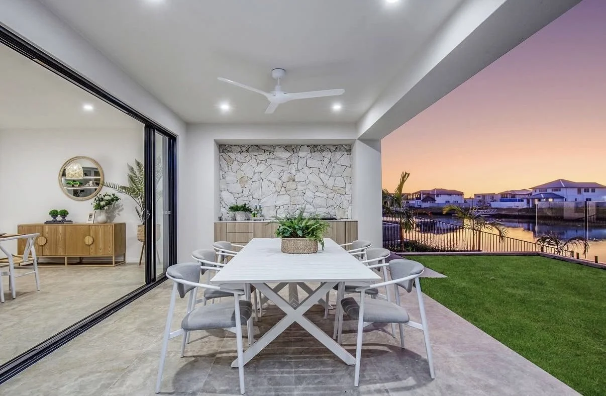 Outdoor patio with a white dining table and chairs, overlooking a body of water and houses at sunset, with a green lawn and glass railing.