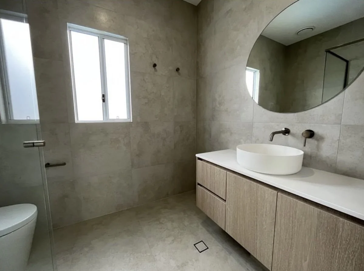 Modern bathroom with a beige tiled wall and floor, a floating wooden vanity with a white oval vessel sink, wall-mounted faucet, a large round mirror, a skylight, and a small window.