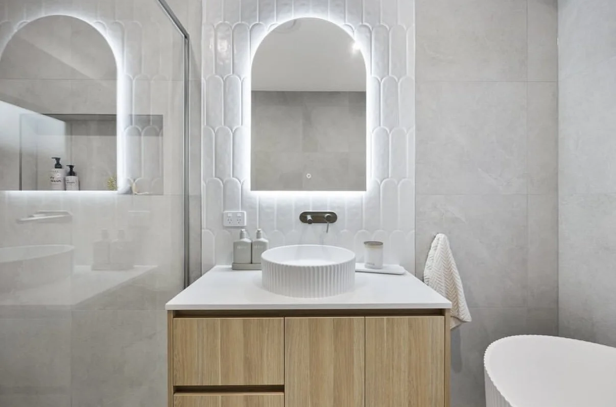 Modern bathroom with a white sink, wooden cabinet, and backlit mirror. Light-colored tiled walls and a towel hanging on the side.