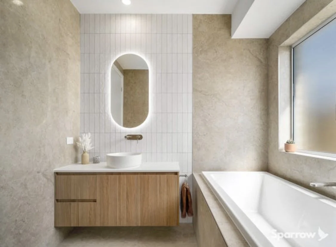 Modern bathroom with a bathtub on the right, a wooden vanity with a vessel sink and an illuminated oval mirror on the left, beige textured walls, and a window with a small potted plant.