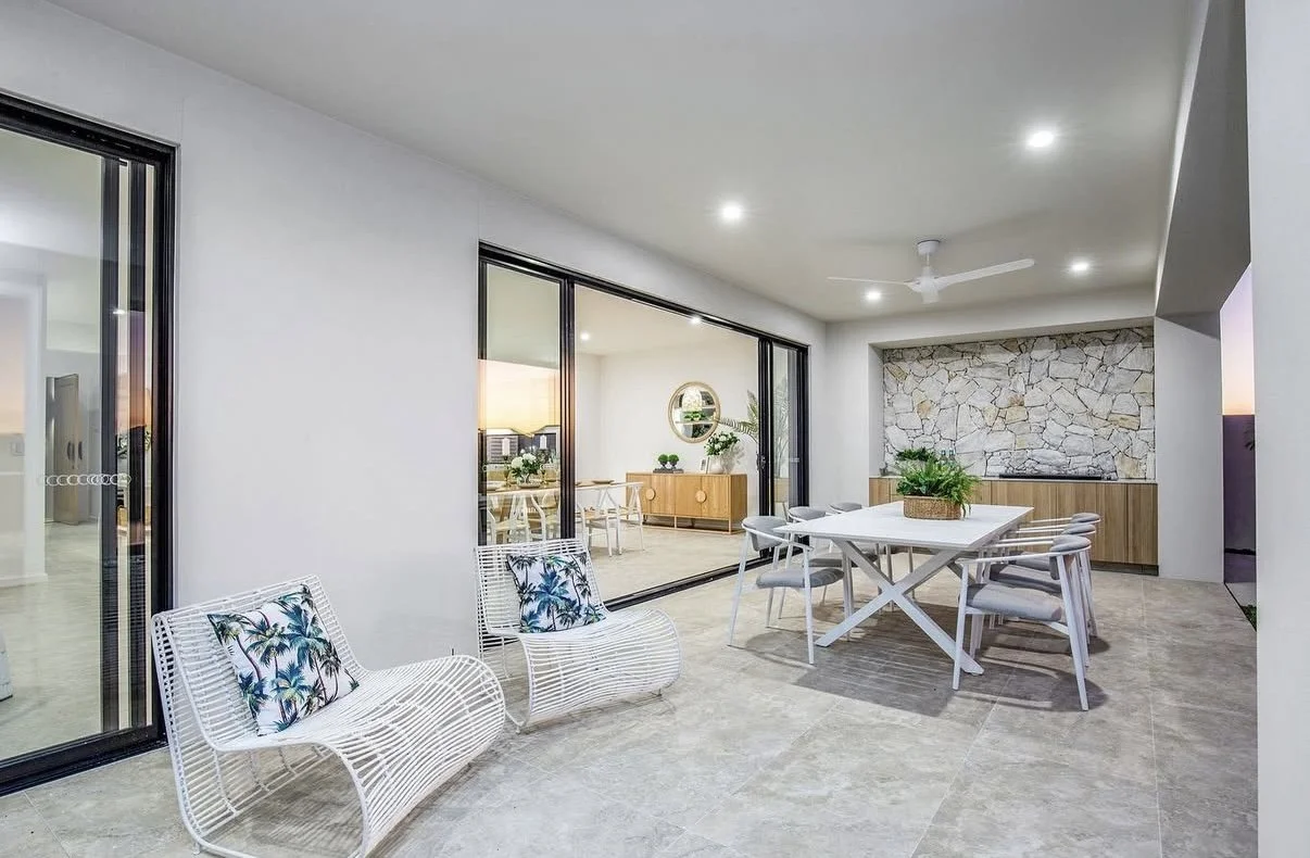 Modern outdoor patio with white wicker chairs with throw pillows, a dining table with six chairs, a stone accent wall, and sliding glass doors leading to an indoor space, set against a sunset sky.