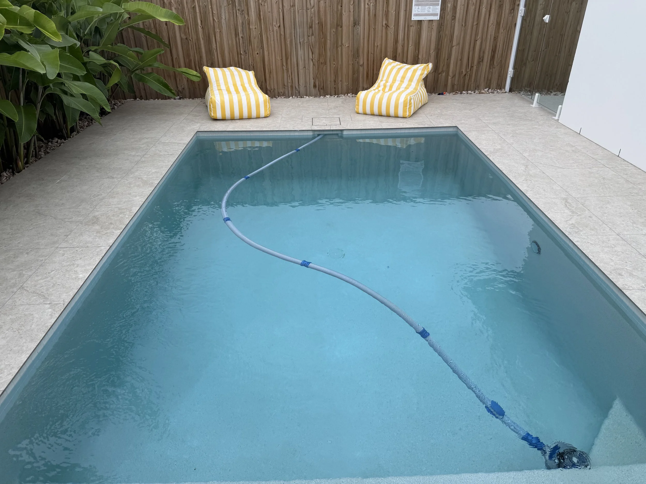 A small rectangular swimming pool with clear water surrounded by a tiled deck. Two yellow and white striped cushioned lounge chairs are placed on the deck, facing the pool. There is a wooden fence in the background, along with a green leafy plant on 