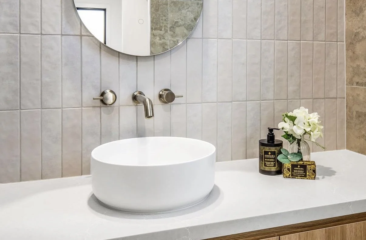 Modern bathroom sink with a round white vessel sink, wall-mounted faucet, and a mirror. Decor includes a black soap pump, a small vase with white flowers, and a bar of soap, all on a white countertop.