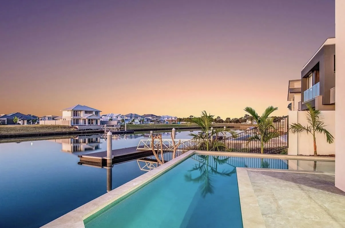 Scenic view of a modern house with a pool overlooking a canal during sunset, with neighboring houses in the background and palm trees near the pool.