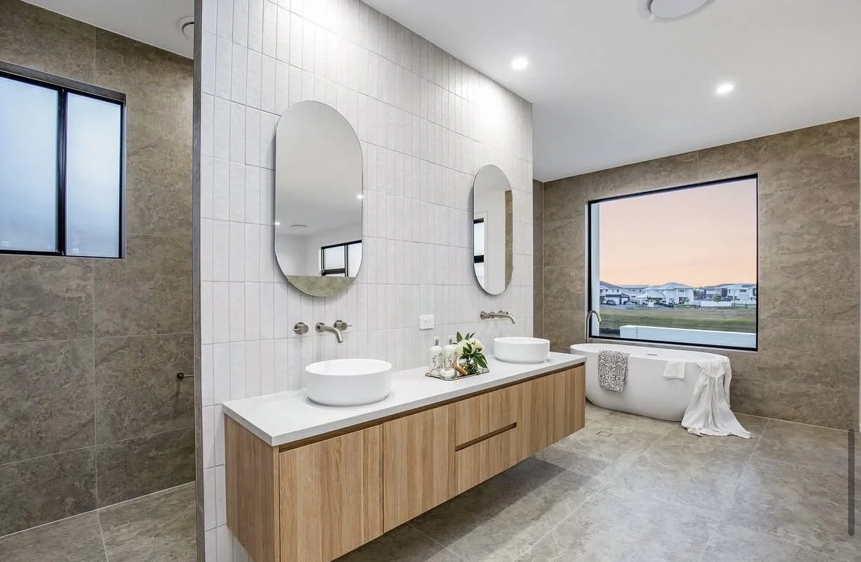 Modern bathroom with double vanity featuring two vessel sinks, large mirrors, beige tiled walls, a freestanding bathtub near a large window, and gray tiled floors.