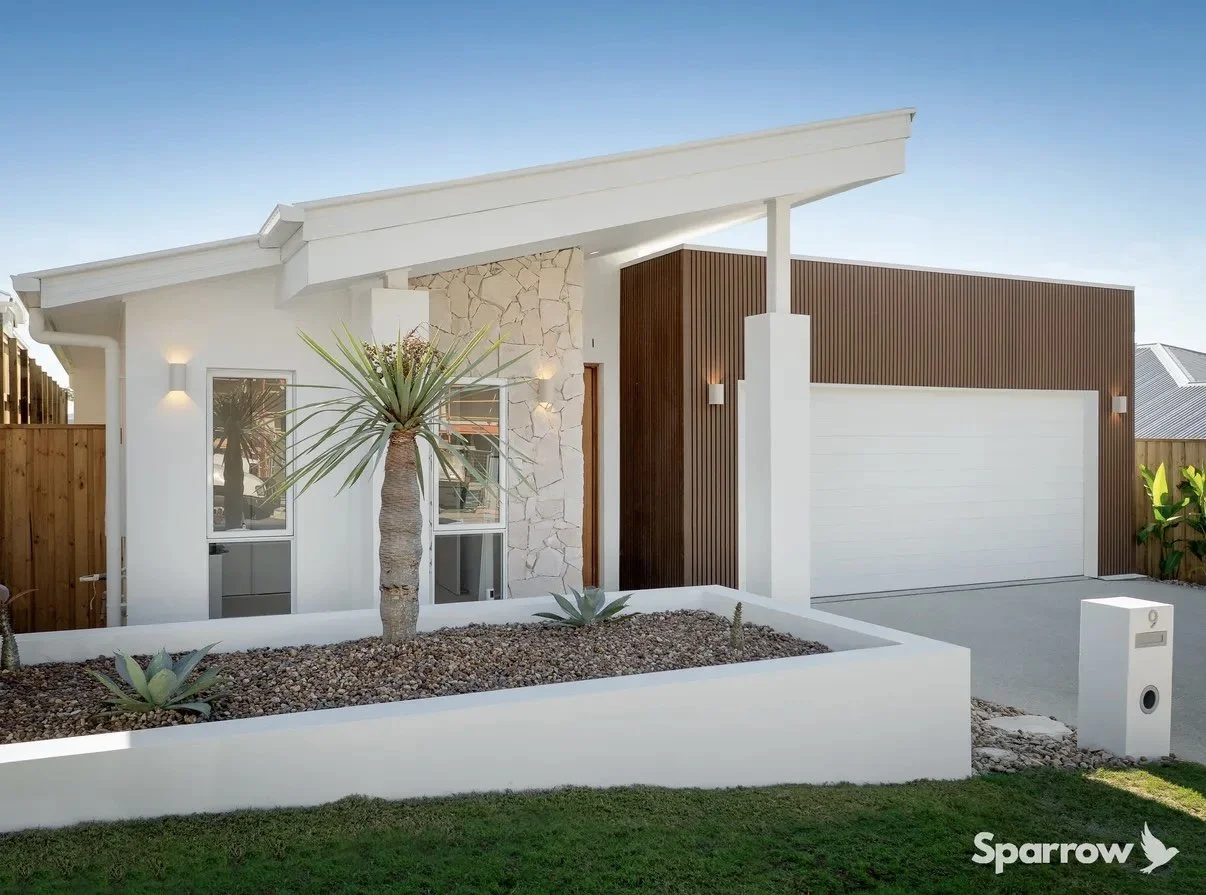 Modern house with white exterior, stone accent wall, and wooden siding. Front yard features desert landscaping with succulents and a tall palm tree. Clear sky in background.