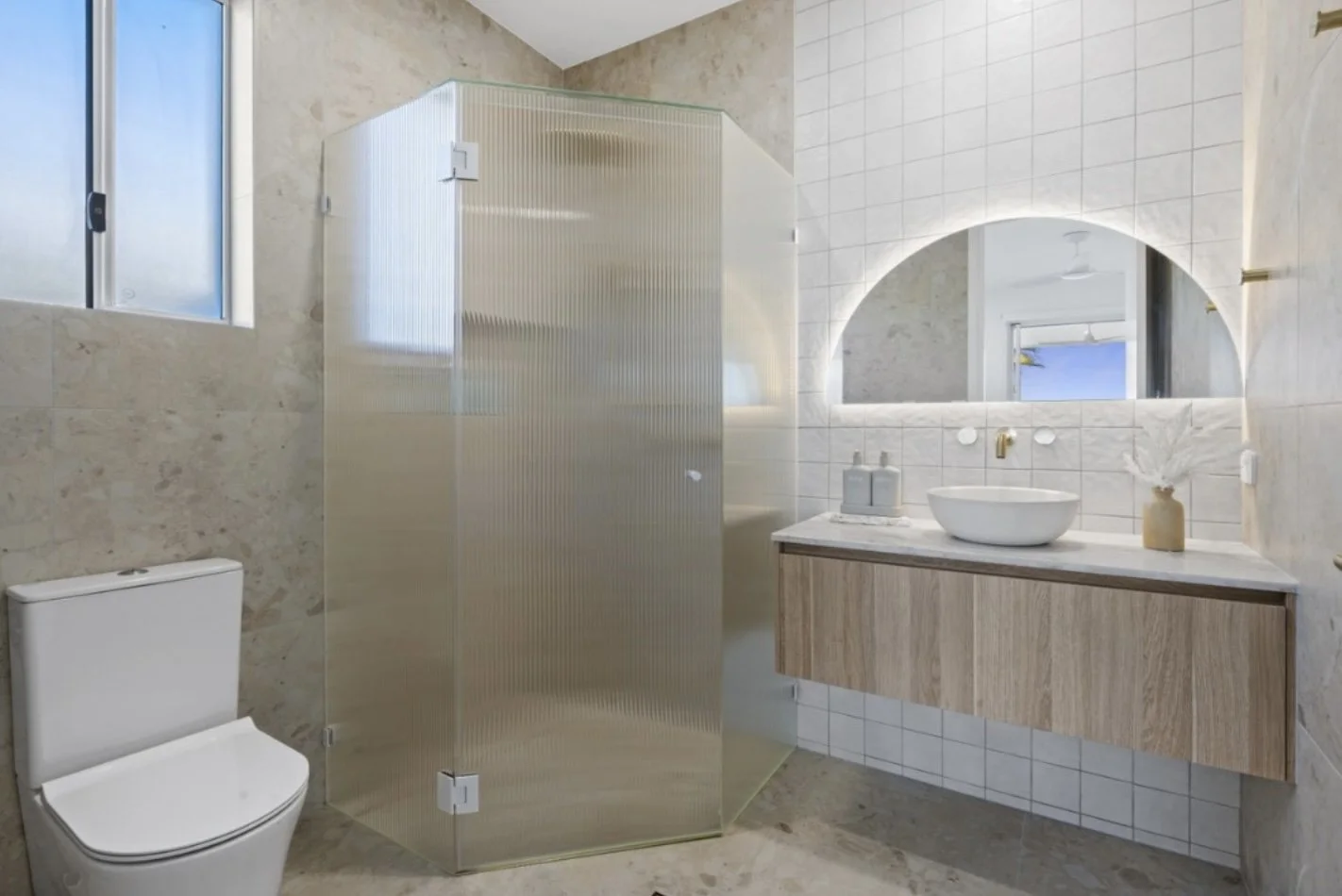 Modern bathroom with a toilet, a frosted shower enclosure, a wooden vanity with a vessel sink, a large mirror with an arch, and a decorative vase with dried flowers.