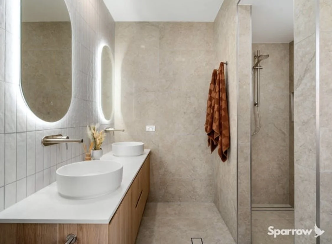 Modern bathroom with double sinks, large mirrors with backlit lighting, beige tiled walls, and a walk-in shower with a brown towel hanging on the wall.