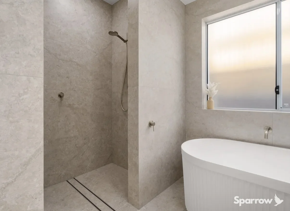 Modern bathroom with a walk-in shower featuring beige tiles and a handheld showerhead. A freestanding bathtub is next to a large frosted window with a decorative vase on the windowsill.