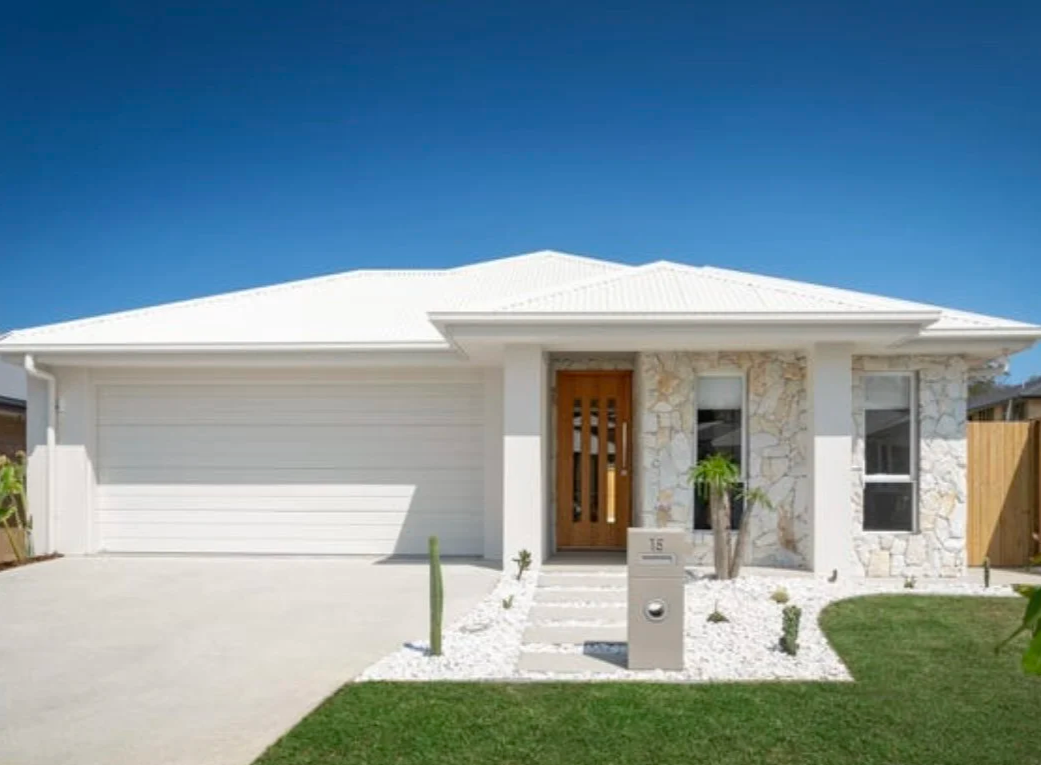 White modern house with stone accents, wooden front door, two windows, a lawn, and a blue sky.
