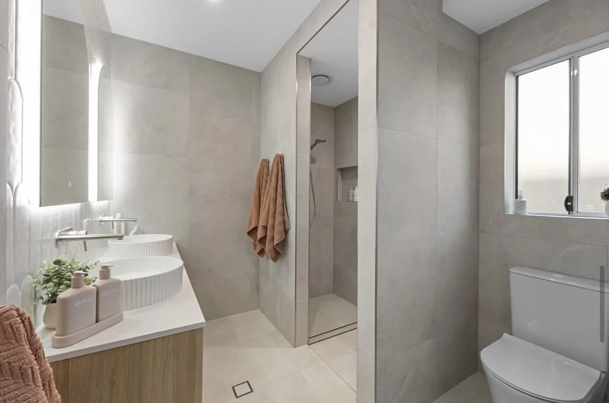 Modern bathroom with a vanity, mirror, walk-in shower, and toilet, featuring beige tiled walls and a window.
